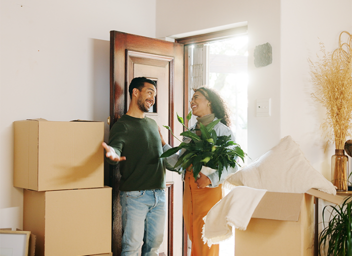 Un hombre y una mujer sonríen entre cajas de cartón en la entrada de una casa.