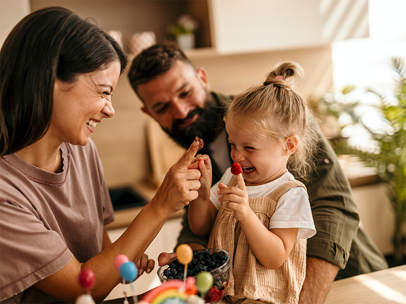 Adultos y niña pequeña ríen jugando con fruta en la mesa.