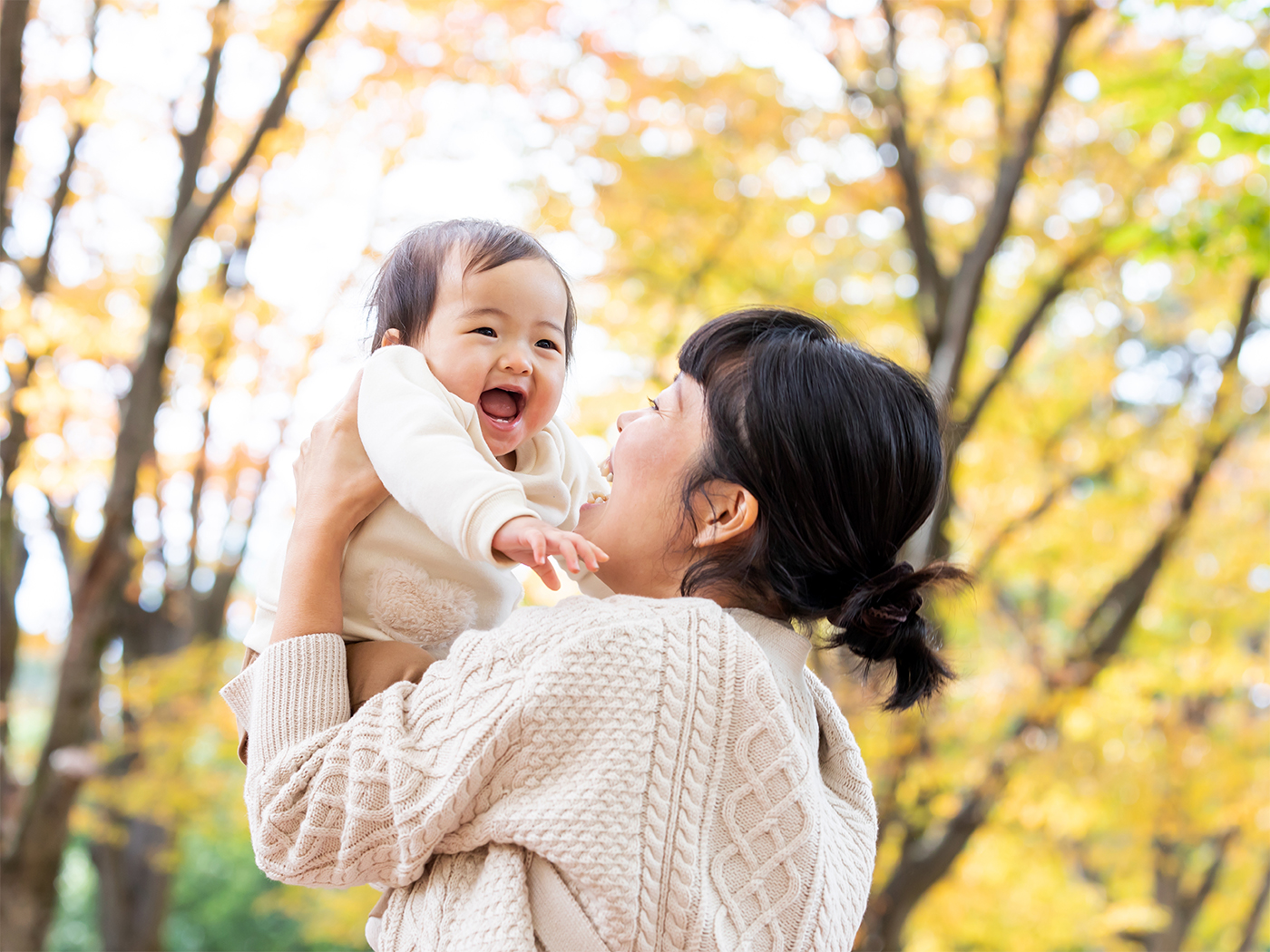 Mujer levantando a un bebé sonriente entre árboles otoñales.