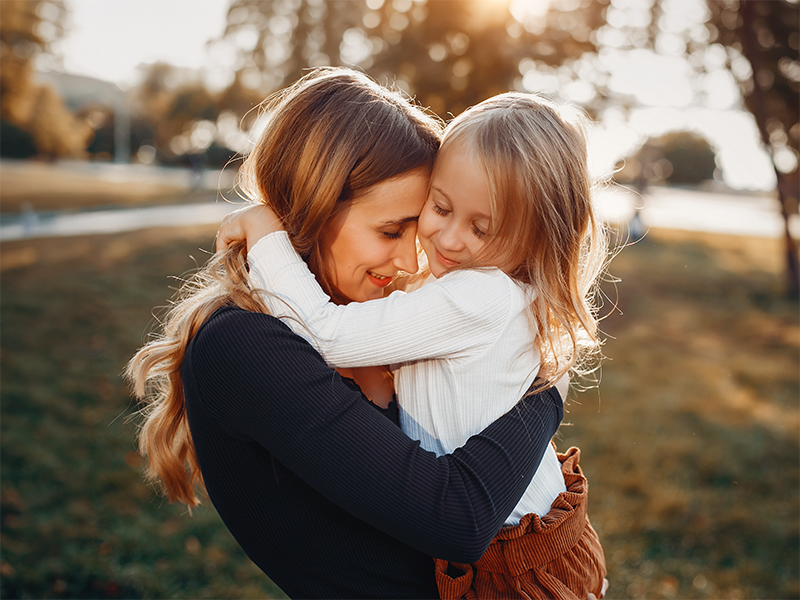 Mujer abraza a una niña en un parque al atardecer.