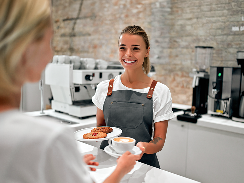 Mujer con delantal sirviendo un café y galletas a otra persona en la barra de una cafetería.