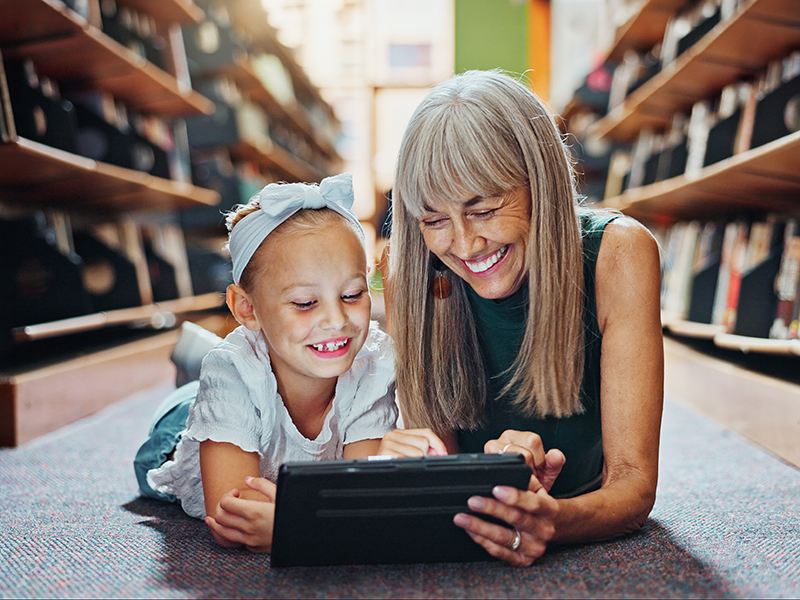 Mujer mayor y niña tumbadas en el suelo de una biblioteca, sonriendo mientras miran una tablet.