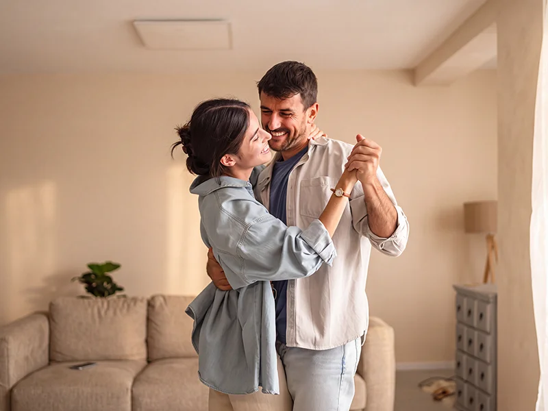 Pareja joven sonriendo mientras bailan abrazados en el salón de su casa.