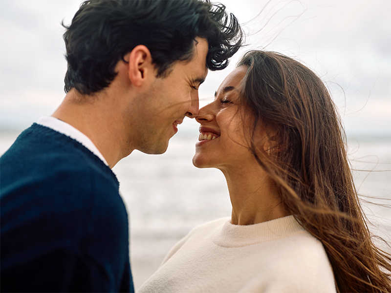 Una pareja sonríe frente con frente junto al mar.