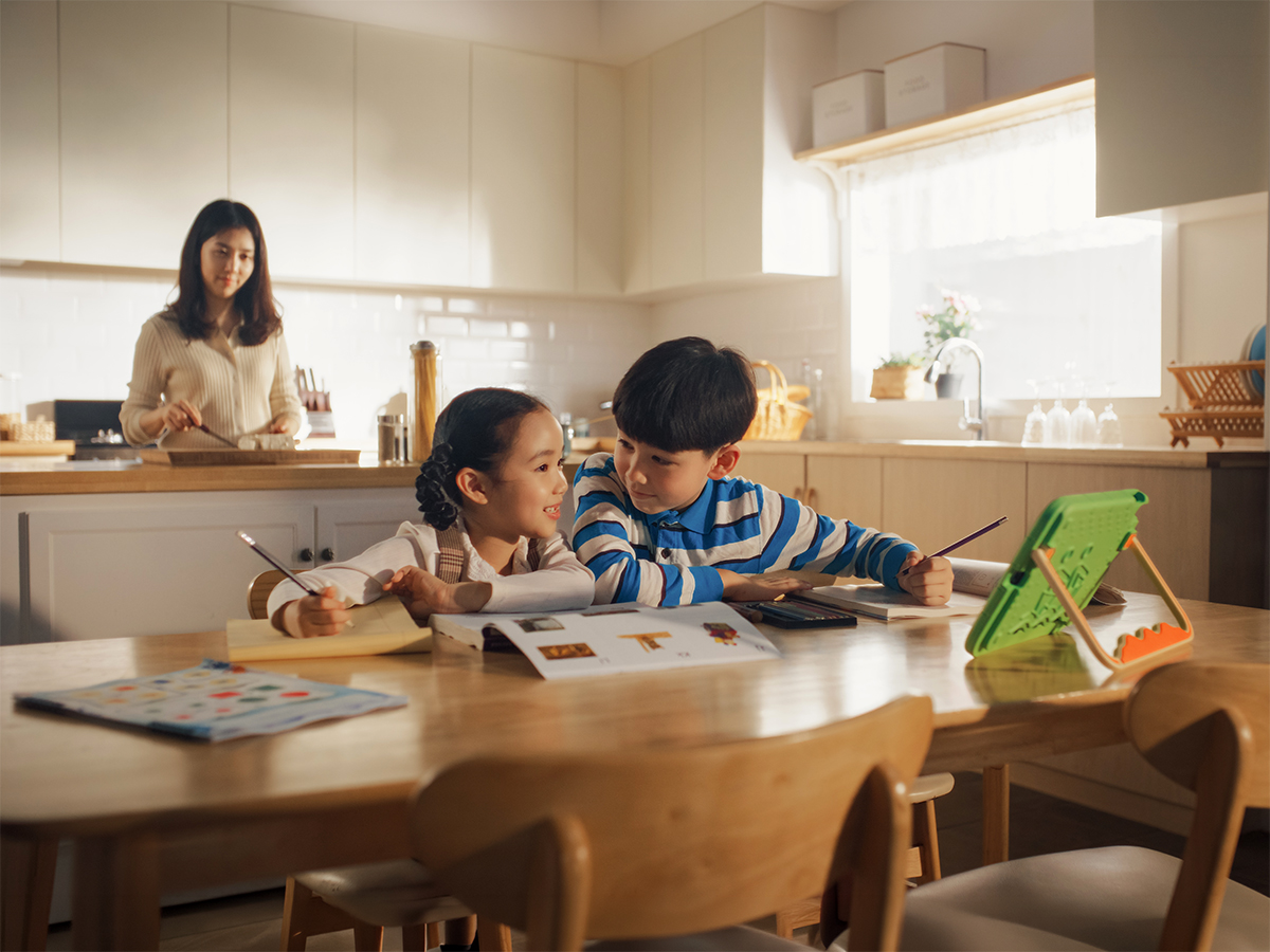 Dos niños haciendo los deberes en la mesa de la cocina mientras una mujer cocina al fondo.