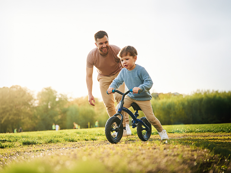 Padre acompaña a su hijo que monta una bicicleta sin pedales en un parque.