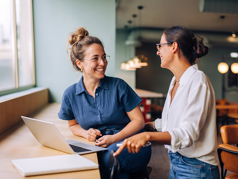 Dos mujeres conversan sonrientes junto a un portátil en una cafetería.