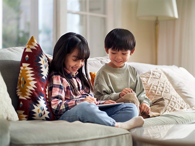 Dos hermanos sonríen mirando una tablet en el sofá.