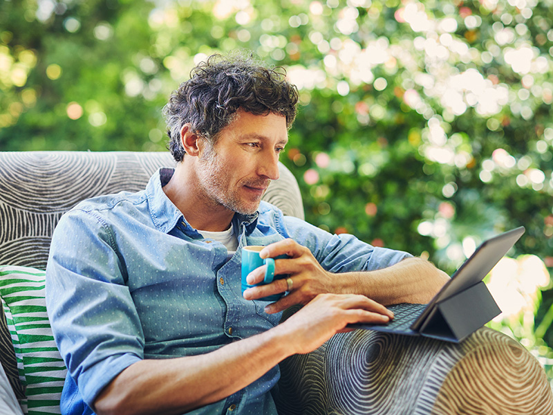 Hombre con una taza usa una tableta en un sillón al aire libre.